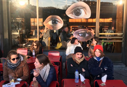 3 kunstwerken in de vorm van ogen op een raam voor een terrasje vol mensen