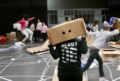 Kinderen rennen door elkaar op podium. Eén jongen heeft een kartonnen doos met twee ogen op het hoofd.