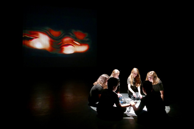 Kinderen in een kring op een donker podium; met achter hen een videoprojectie